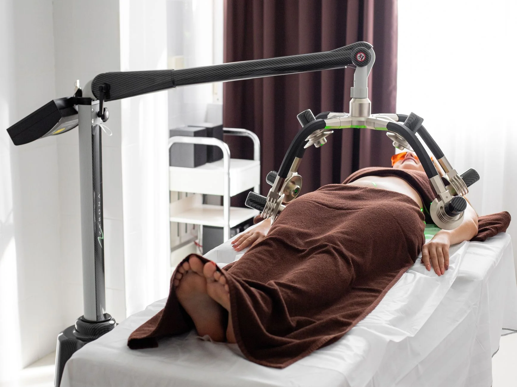 Faceless female patient covered with towels lying under laser fat loss machine during treatment in light medical clinic with curtains on window at House of Aetheria, Gurugram